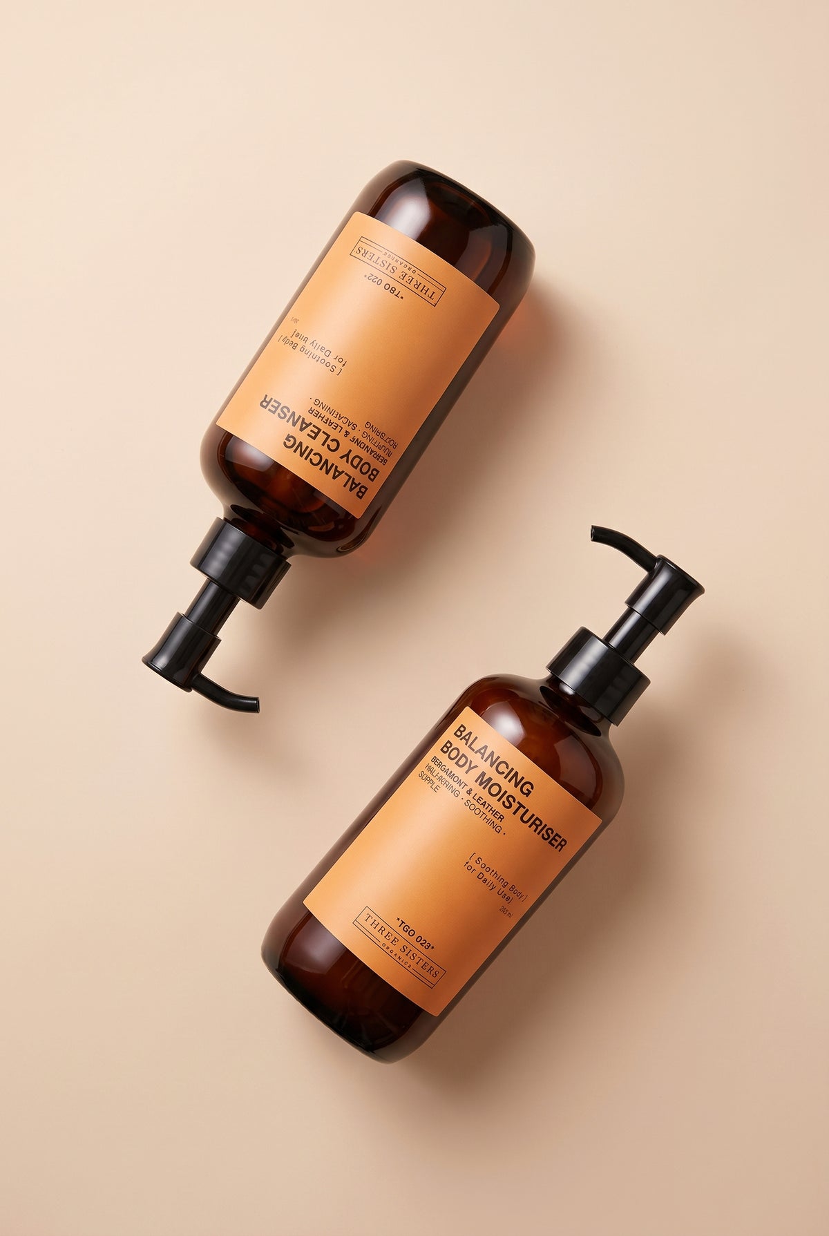 Two amber glass body care bottles with black pumps and tan labels, arranged diagonally on a warm beige background. One bottle is labeled body cleanser and the other body moisturizer.