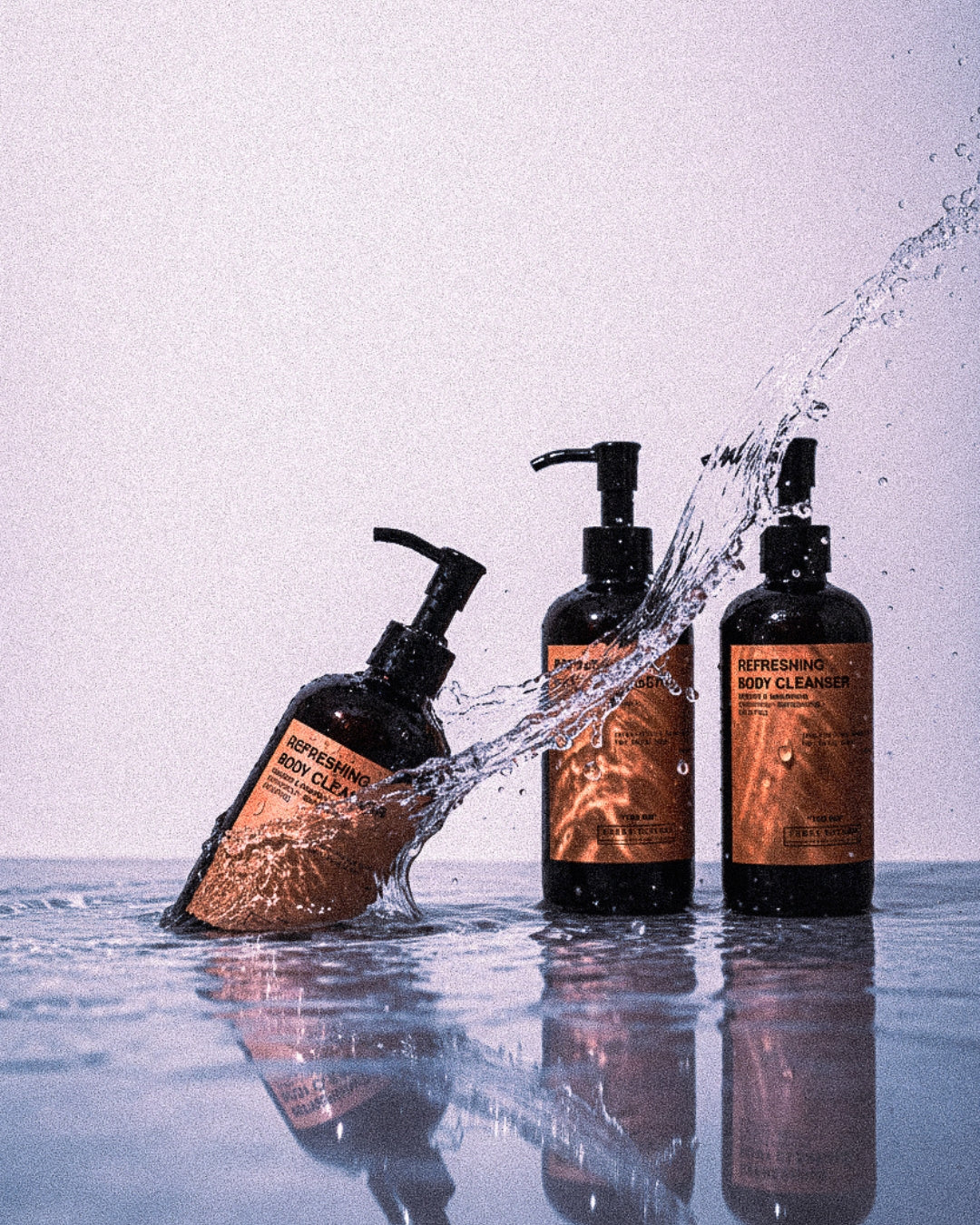 Three bottles of body cleanser with water splashing around them on a reflective surface.